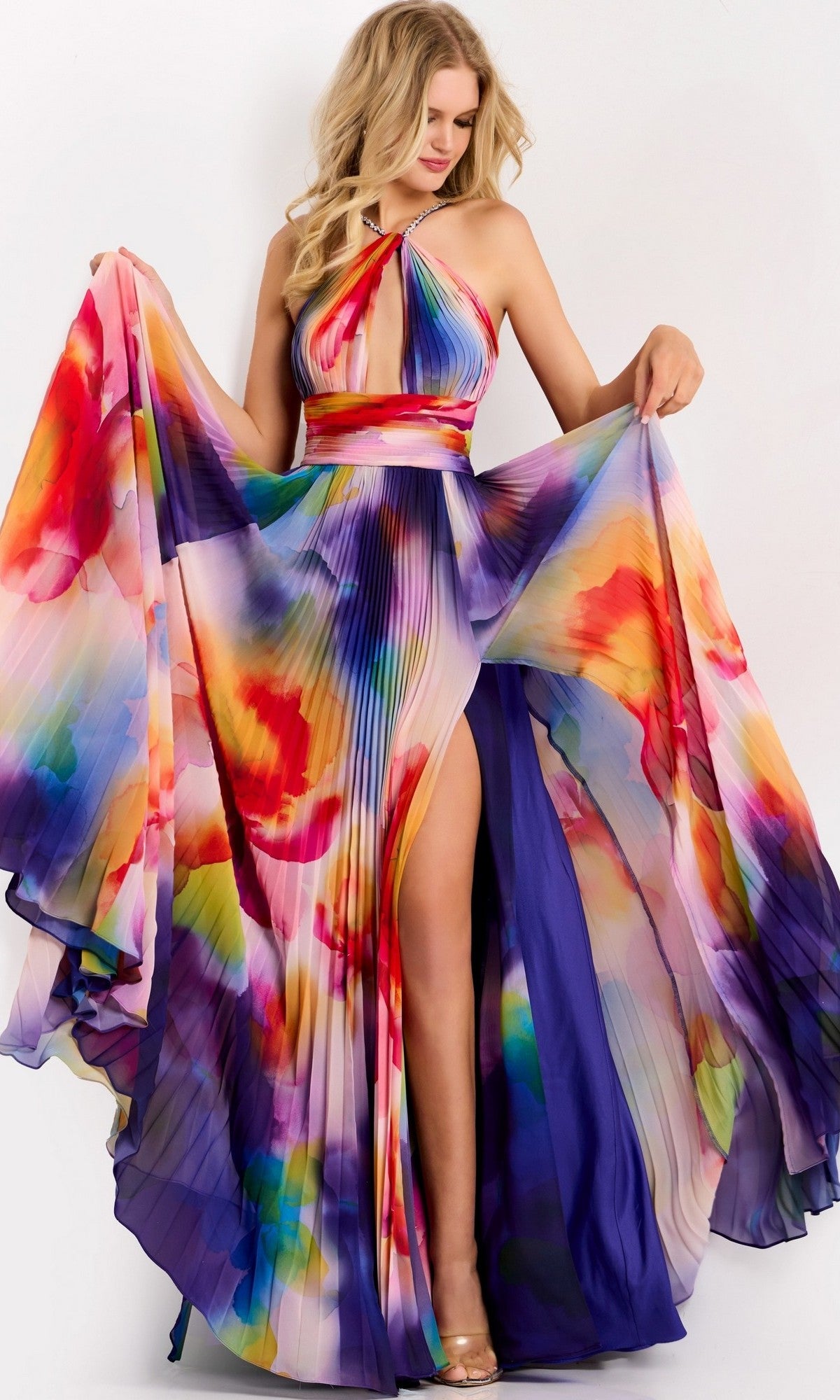 A woman poses in the Jovani 42717 Long Prom Dress, a colorful pleated gown with a halter neckline and high slit, featuring a vibrant watercolor pattern in red, orange, yellow, blue, and purple.